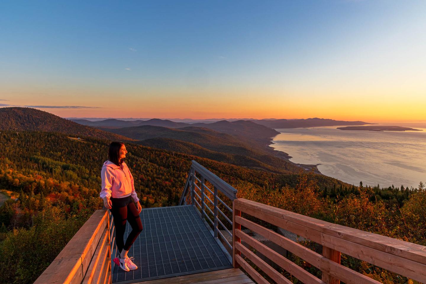 Personne sur une terrasse observant un coucher de soleil sur un paysage de montagne et de mer.