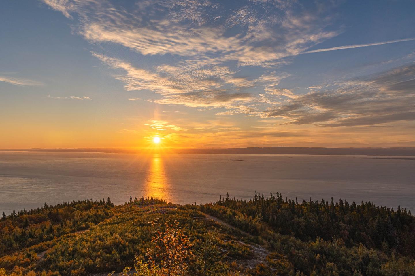 Coucher de soleil sur la mer, ciel nuageux, colline verdoyante au premier plan.