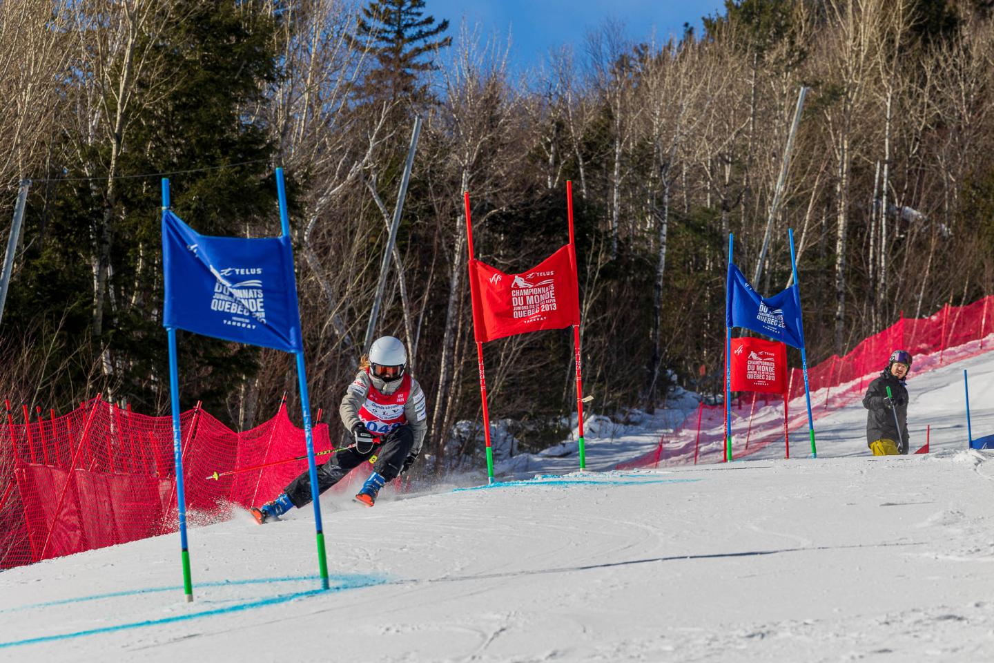 Skieur slalomant entre des portes colorées, neige, ciel bleu, forêt en arrière-plan.