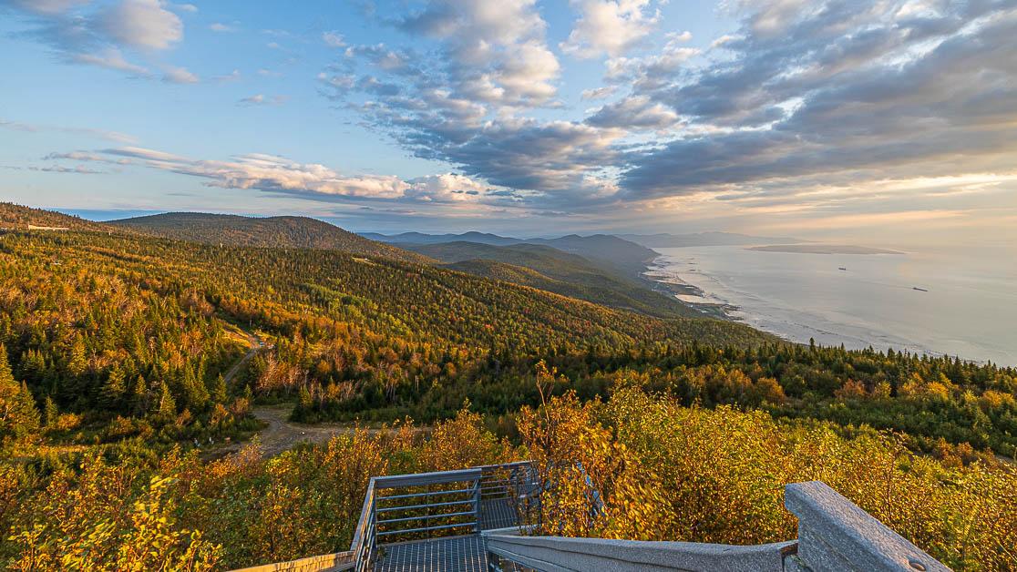 Vue panoramique d'une forêt automnale et mer à l'horizon sous un ciel partiellement nuageux.