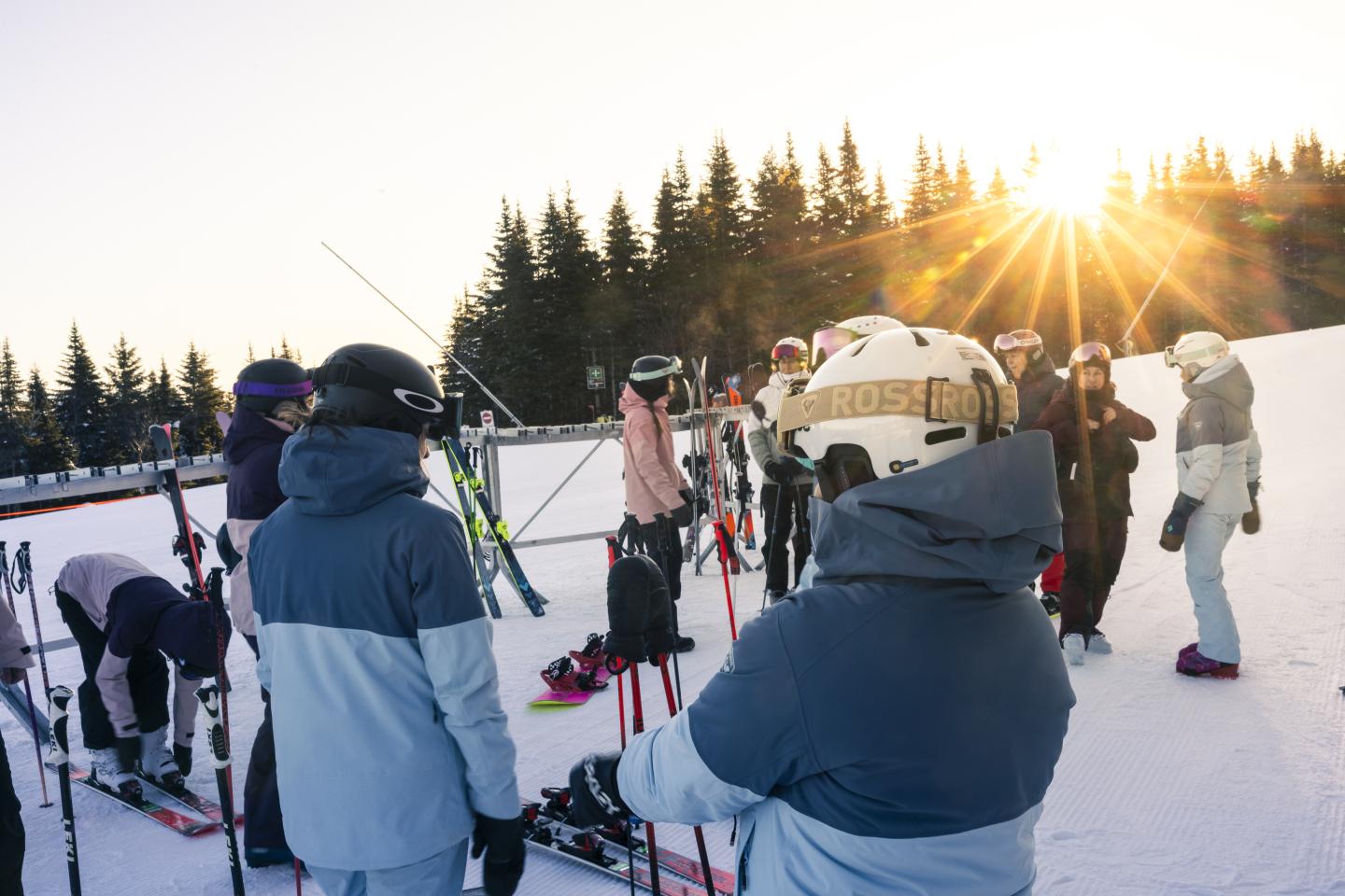Personnes en tenue de ski sur une pente enneigée au coucher de soleil, forêt en arrière-plan.