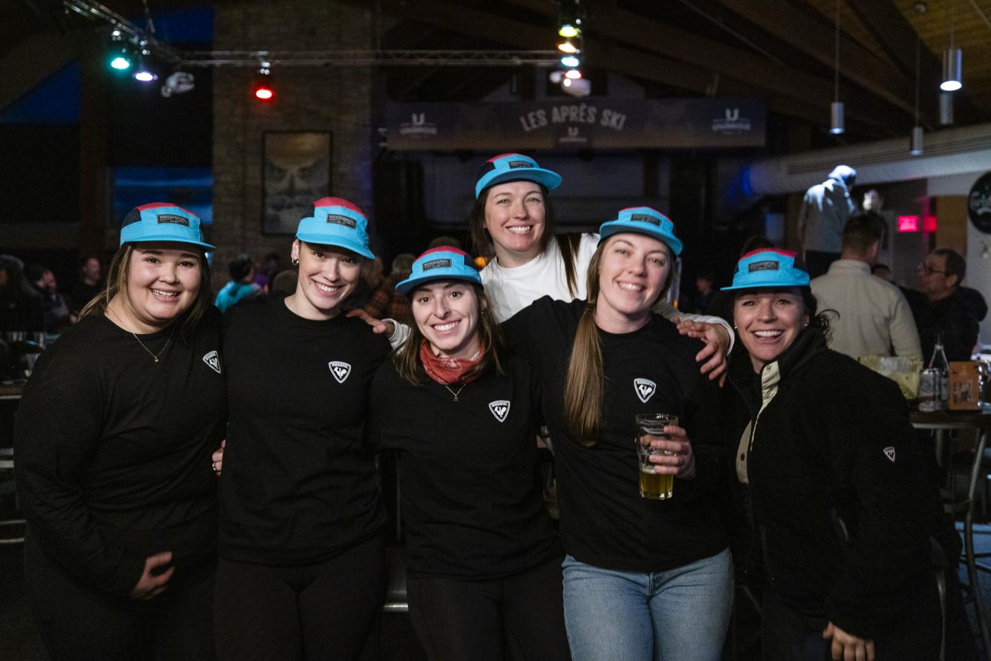 Groupe de femmes souriantes, portant des casquettes bleues, dans un bar animé.
