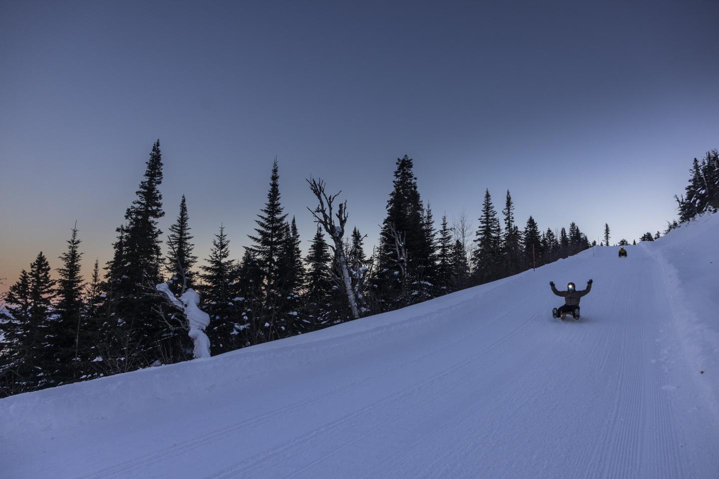 Snowboarder sur une piste enneigée, forêt en arrière-plan, ciel au crépuscule.