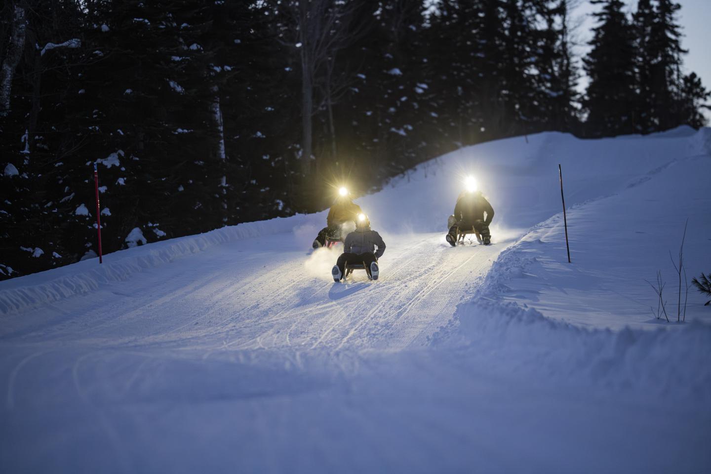 Scooters des neiges illuminant un chemin enneigé crépusculaire entouré d'arbres.