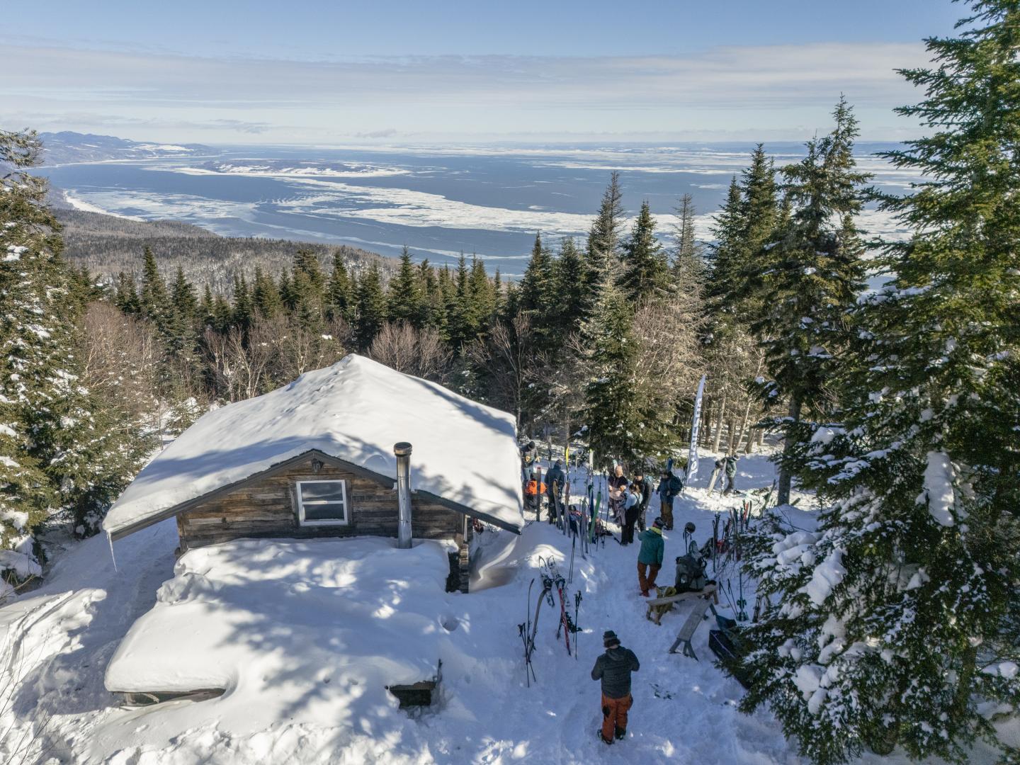 Chalet enneigé entouré de skieurs et de sapins, vue sur mer et montagnes.