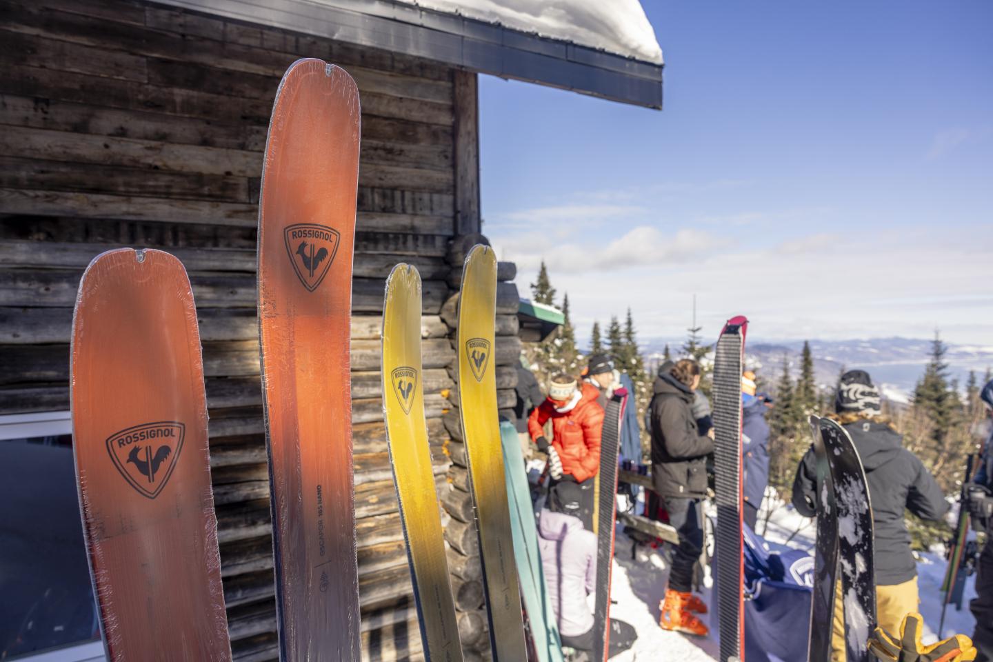 Skis colorés appuyés contre un chalet enneigé, skieurs en arrière-plan.