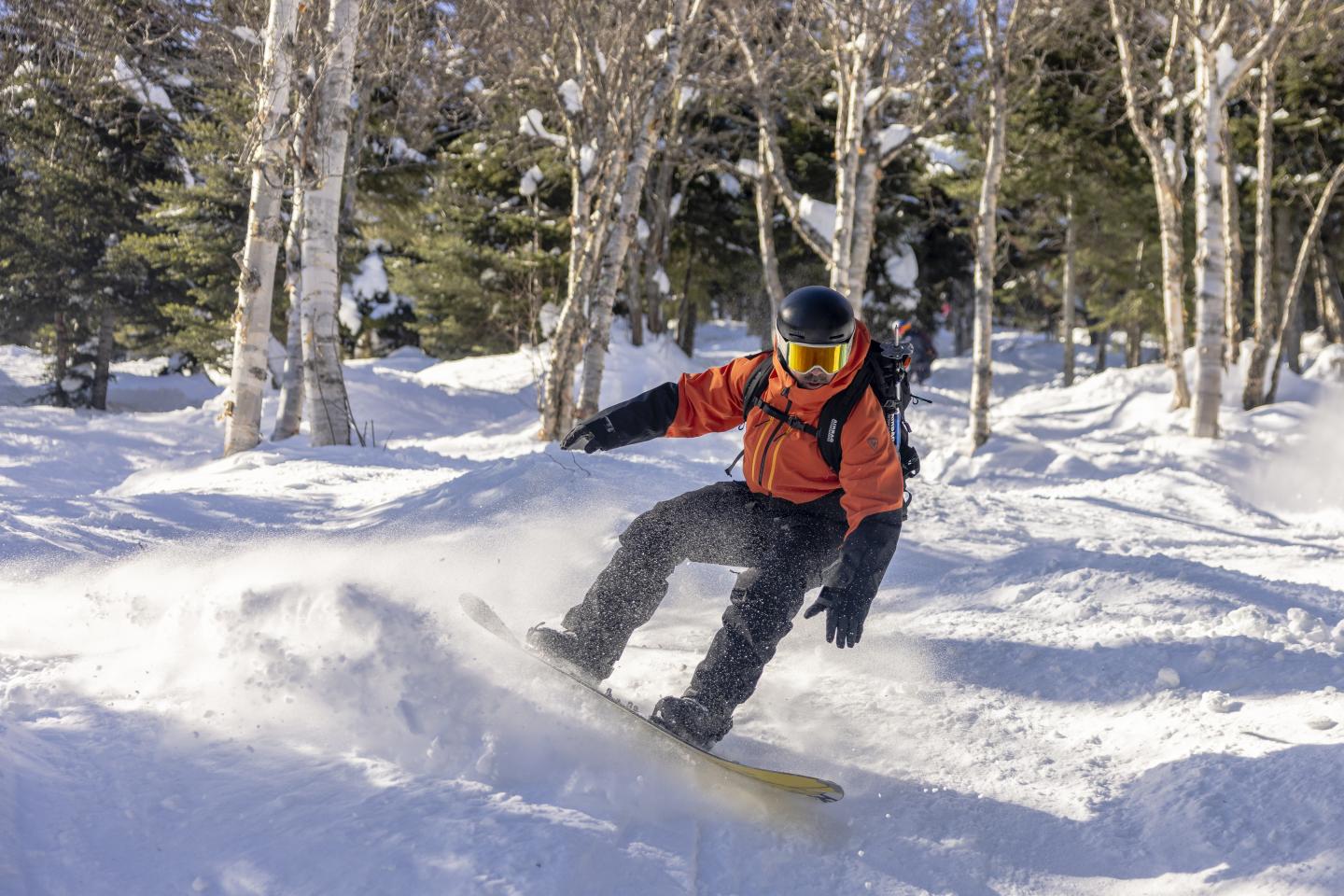 Snowboardeur en plein mouvement dans une forêt enneigée, portant veste orange et casque noir.