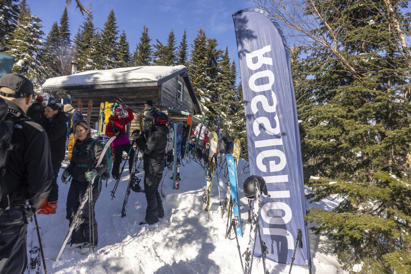 Skieurs rassemblés près d'un chalet enneigé, forêt de sapins en arrière-plan.