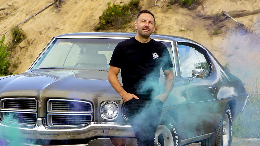 Un homme souriant debout devant une voiture ancienne, avec de la fumée colorée autour.