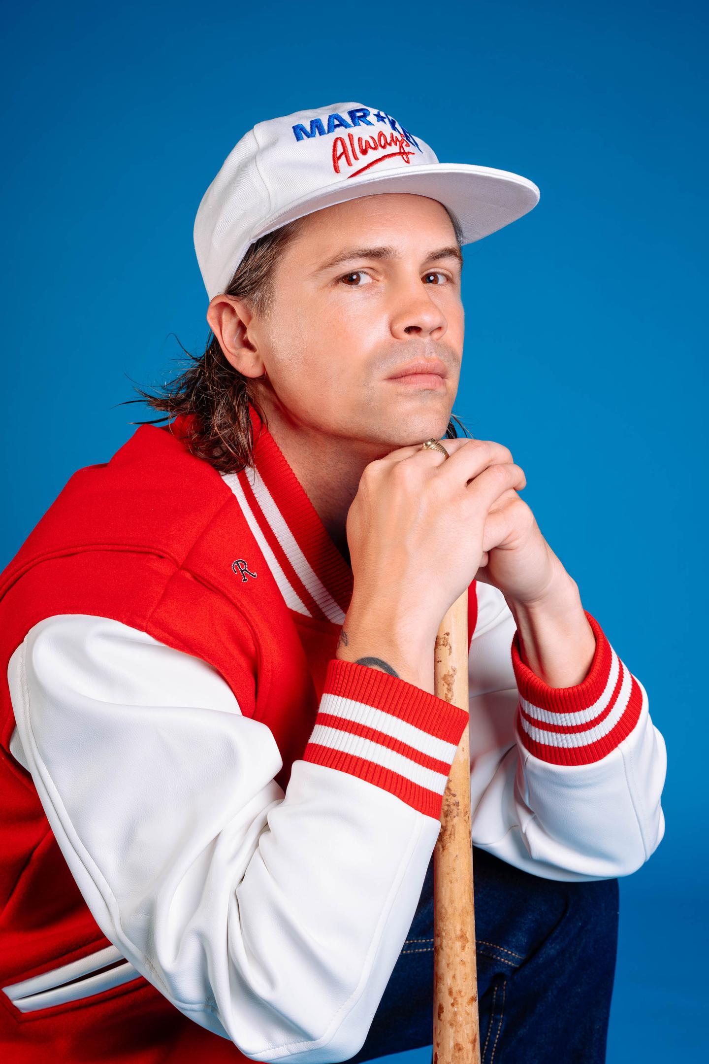 Homme en veste rouge et blanche, casquette, fond bleu.