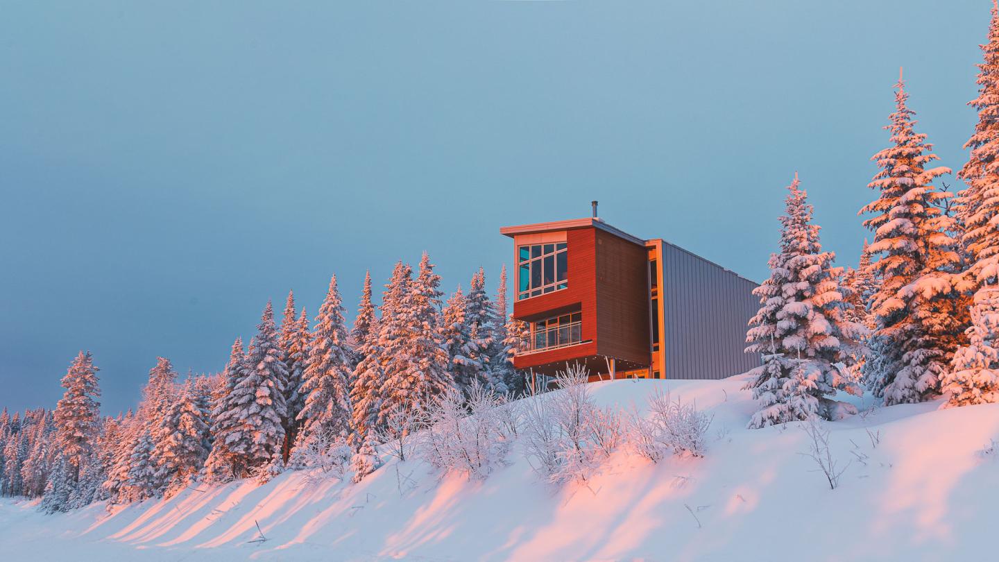 Maison moderne surplombant une colline enneigée, entourée de sapins sous un ciel bleu.