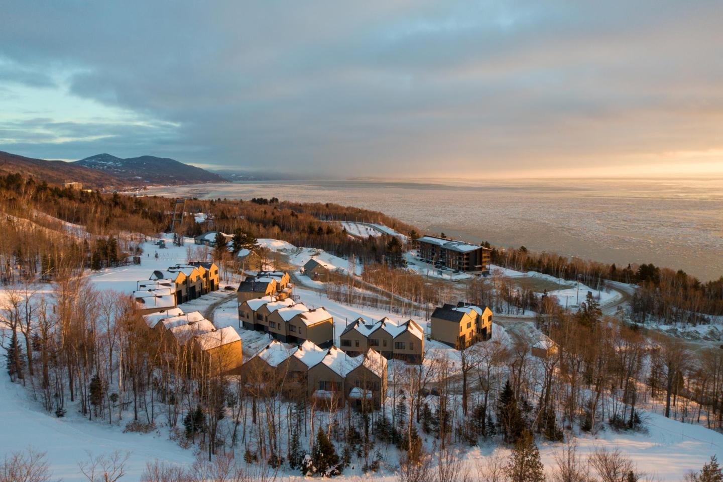 Village enneigé au lever du soleil, vue aérienne avec mer et montagnes à l'horizon.