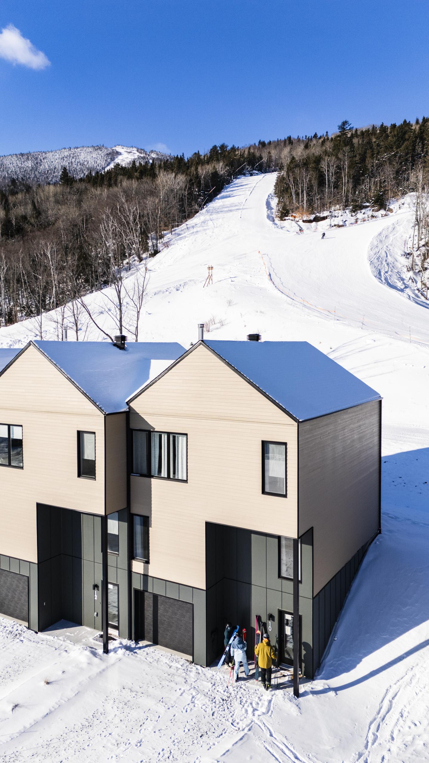 Deux maisons modernes près d'une piste enneigée sous un ciel bleu.