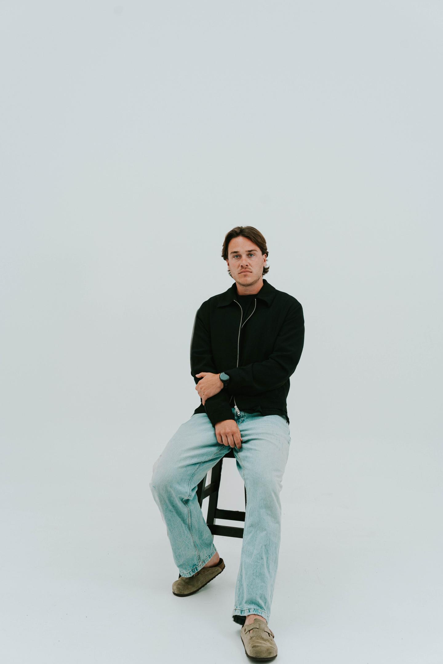 Homme assis sur un tabouret dans un studio au fond clair.