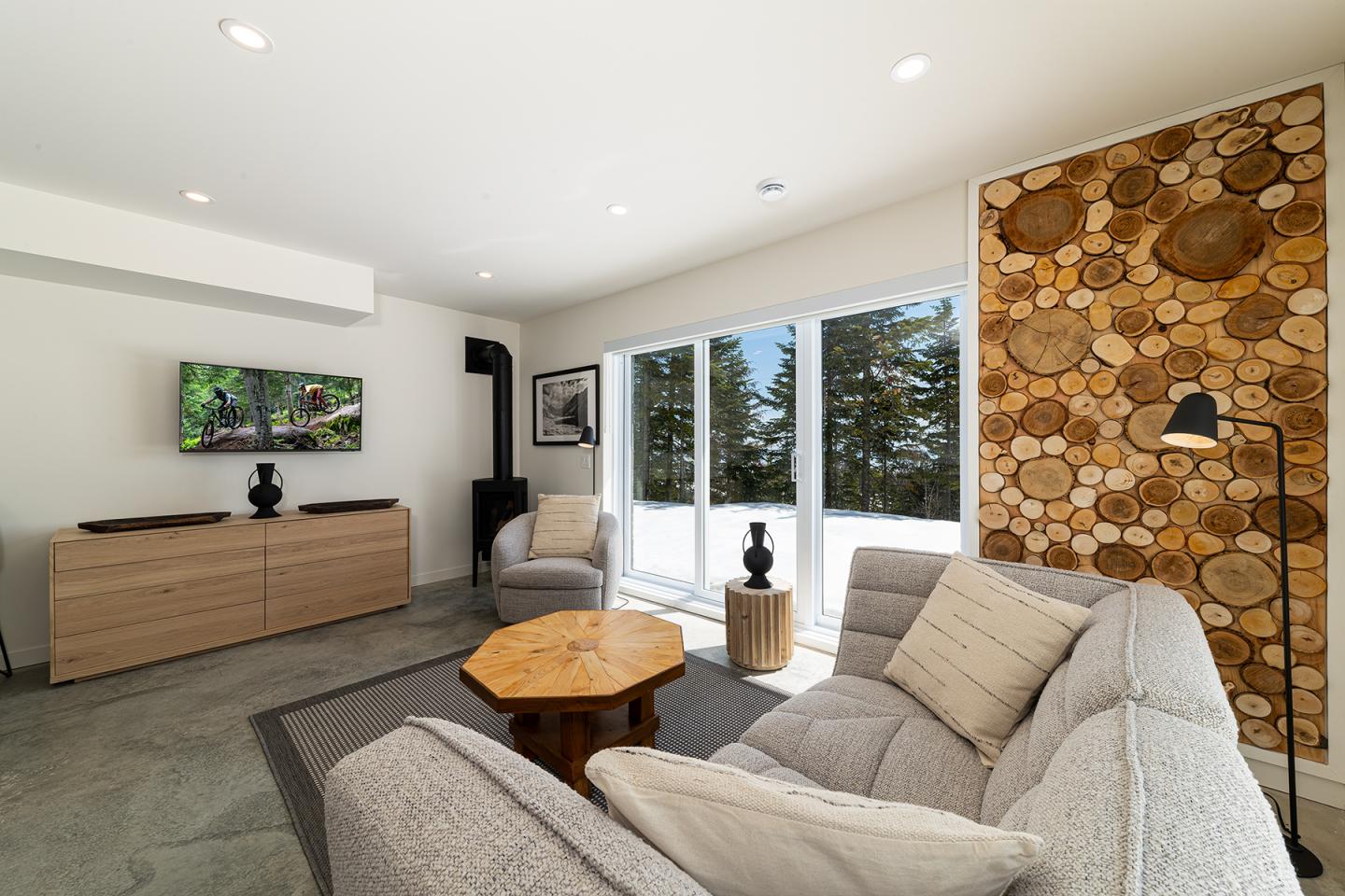Salon moderne avec canapé gris, table basse en bois, et baie vitrée sur forêt enneigée.