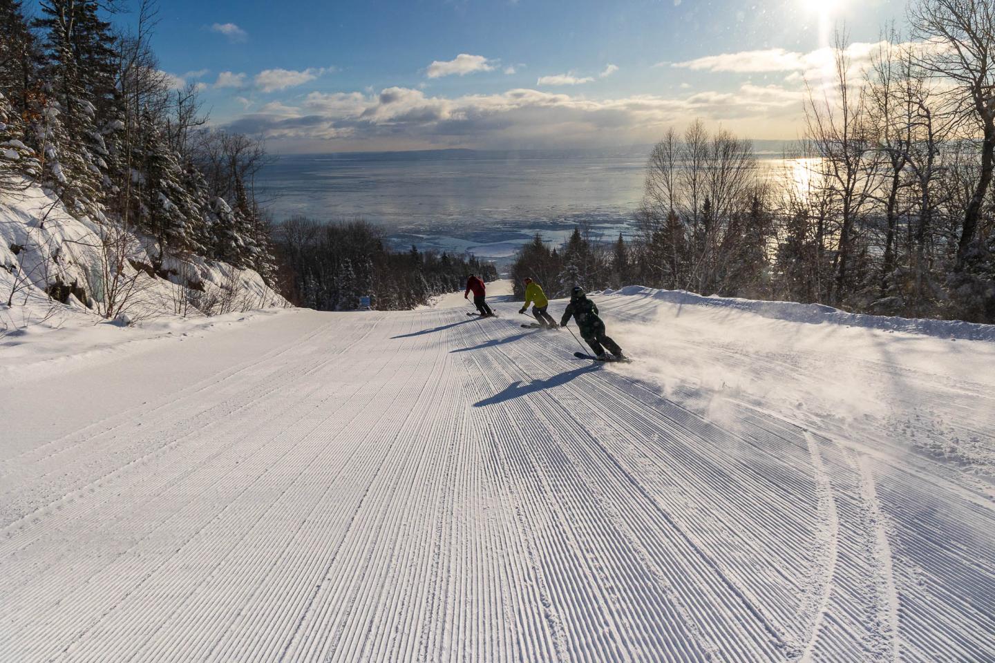 Des skieurs descendent une piste enneigée, vue sur la mer en arrière-plan.