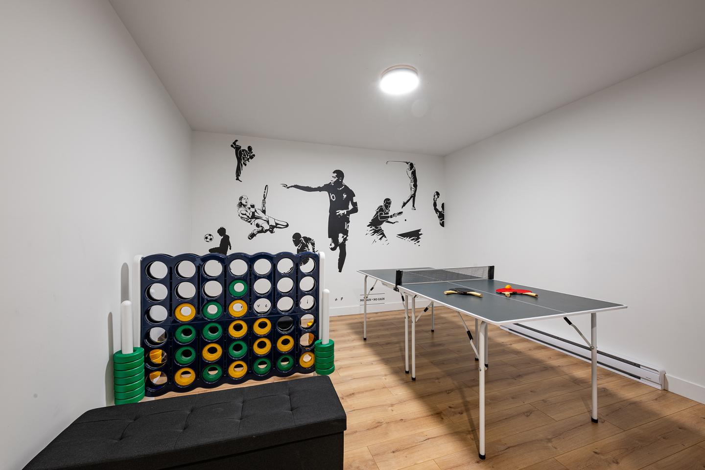 Salle de jeux avec table de ping-pong et jeu Puissance 4 géant, mur décoré de silhouettes sportives.