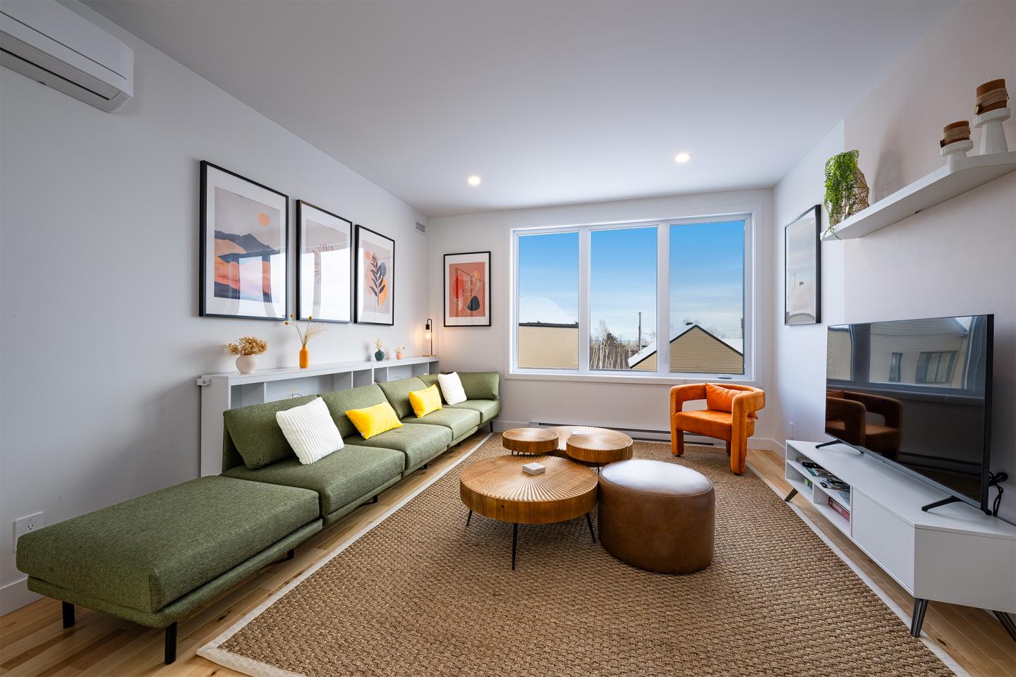 Salon moderne avec canapé vert, tables basses en bois, fauteuil orange et grande fenêtre.