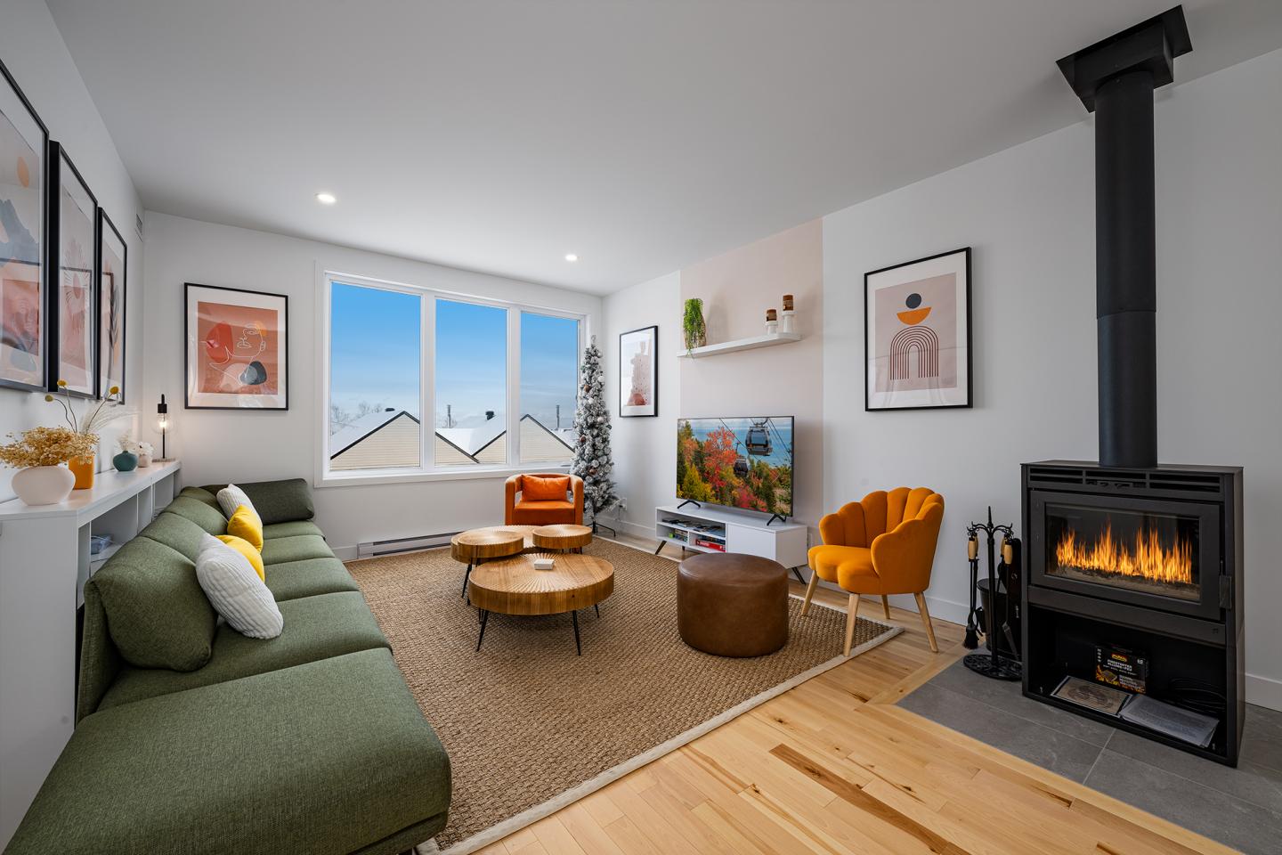 Salon moderne avec canapé vert, cheminée allumée, fauteuils orange et vue sur montagnes.