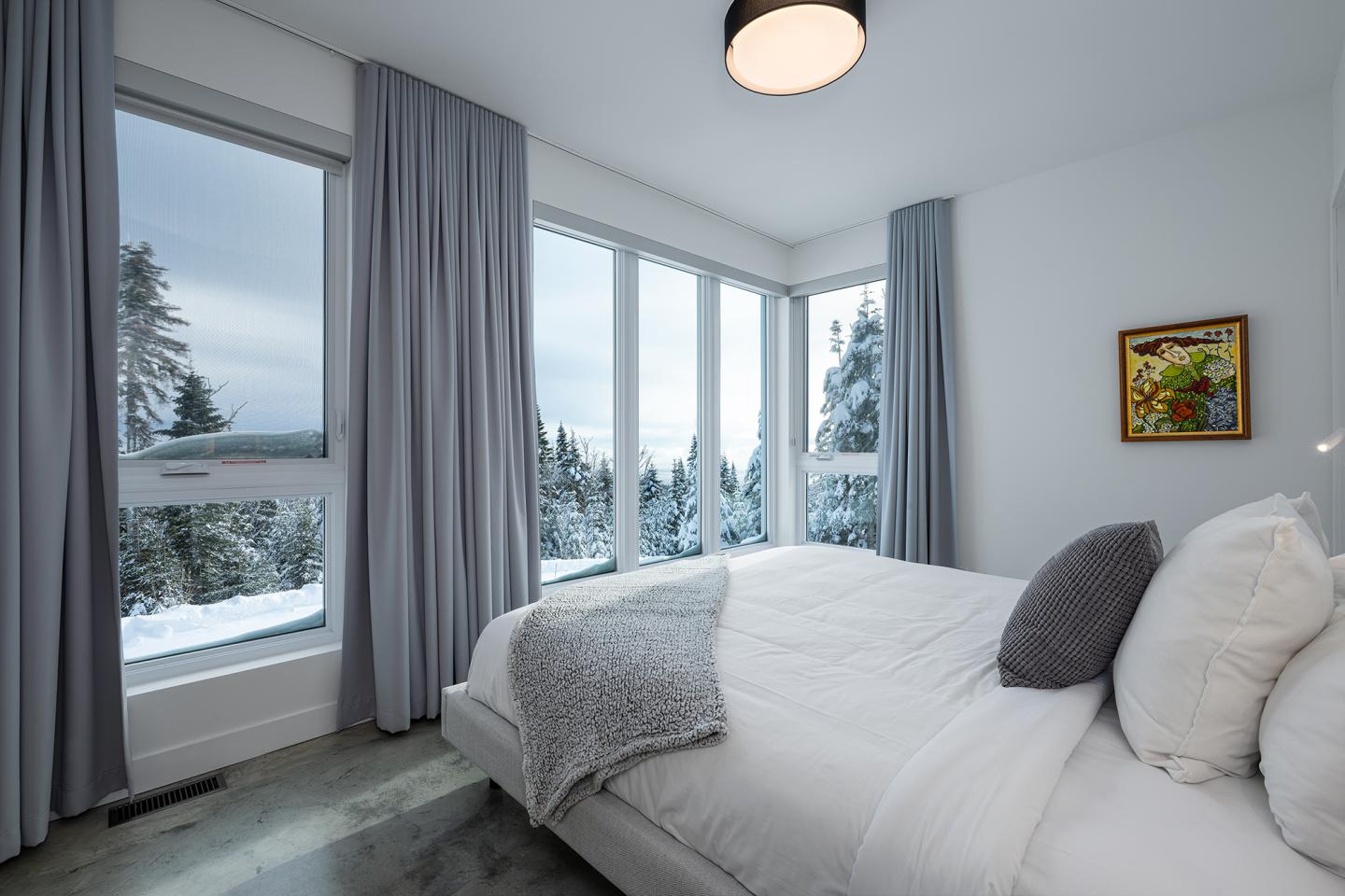 Chambre moderne avec vue sur forêt enneigée, lit blanc et rideaux gris.