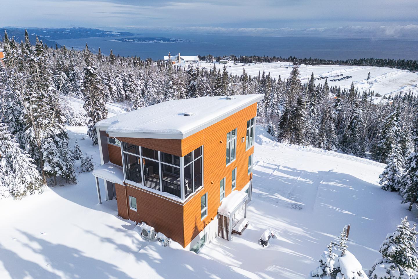 Maison en bois entourée de neige et de sapins, vue sur la mer et montagnes.