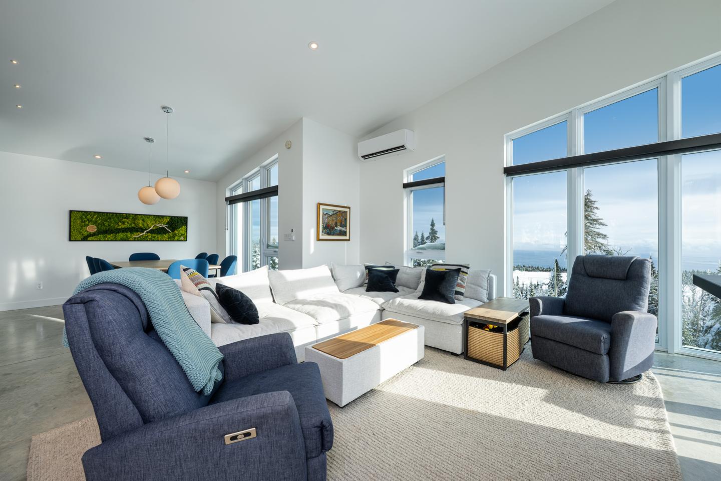 Salon lumineux avec canapés blancs, fauteuils gris, grandes fenêtres et vue sur montagne.