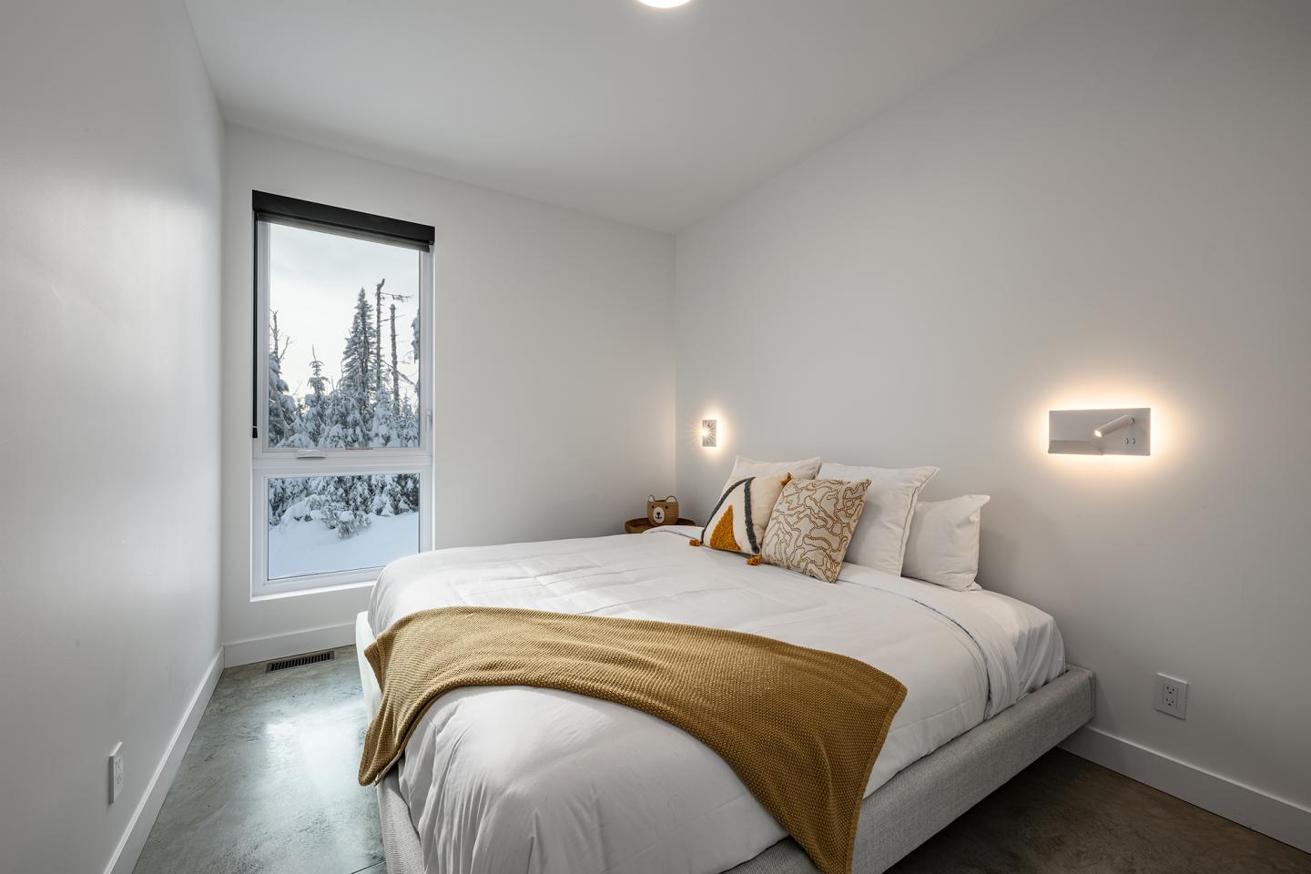 Chambre minimaliste avec lit blanc, plaid jaune, fenêtre sur paysage enneigé.