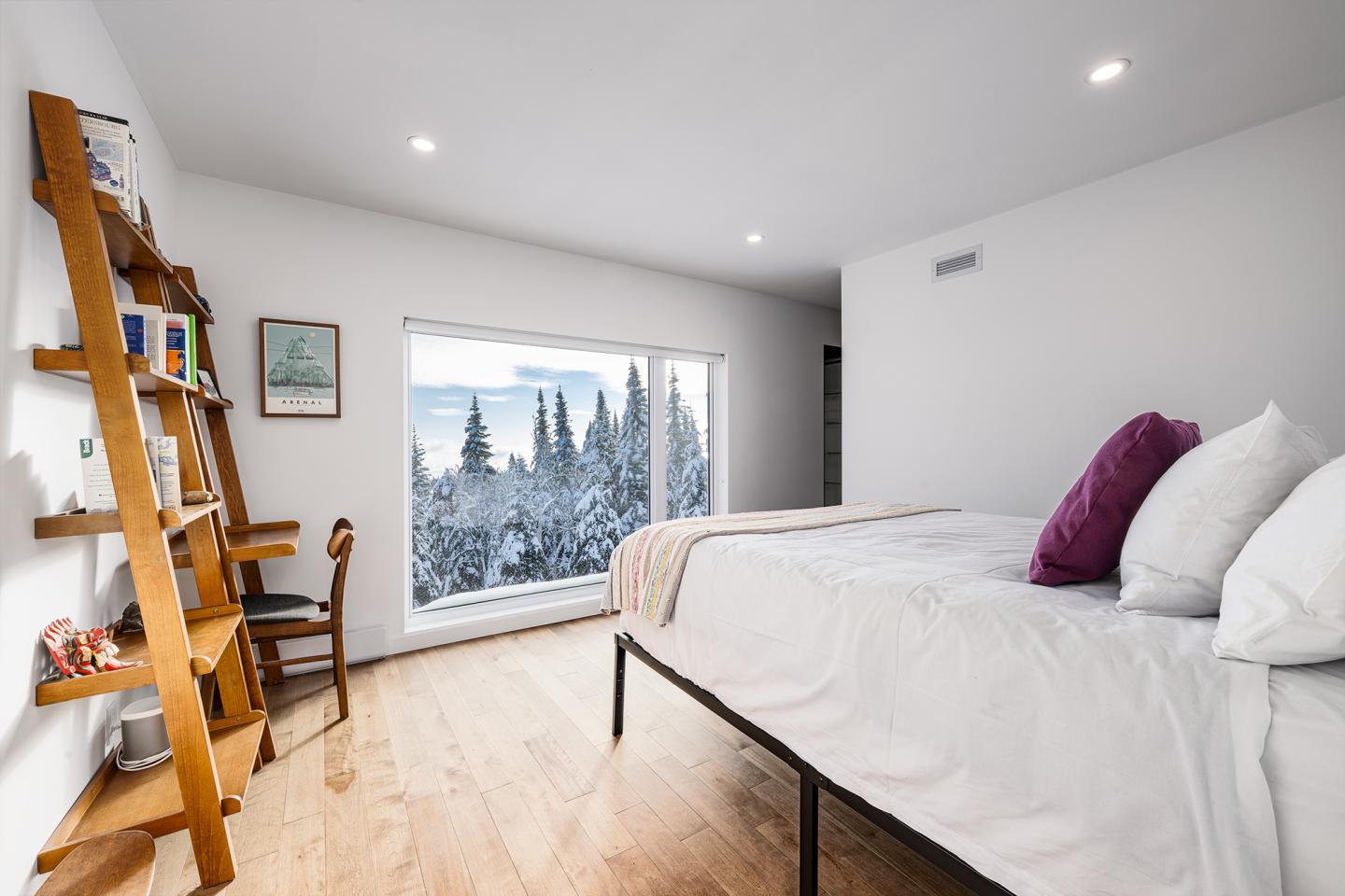 Chambre moderne avec lit blanc et vue sur paysage neigeux. Échelle en bois et chaise à gauche.