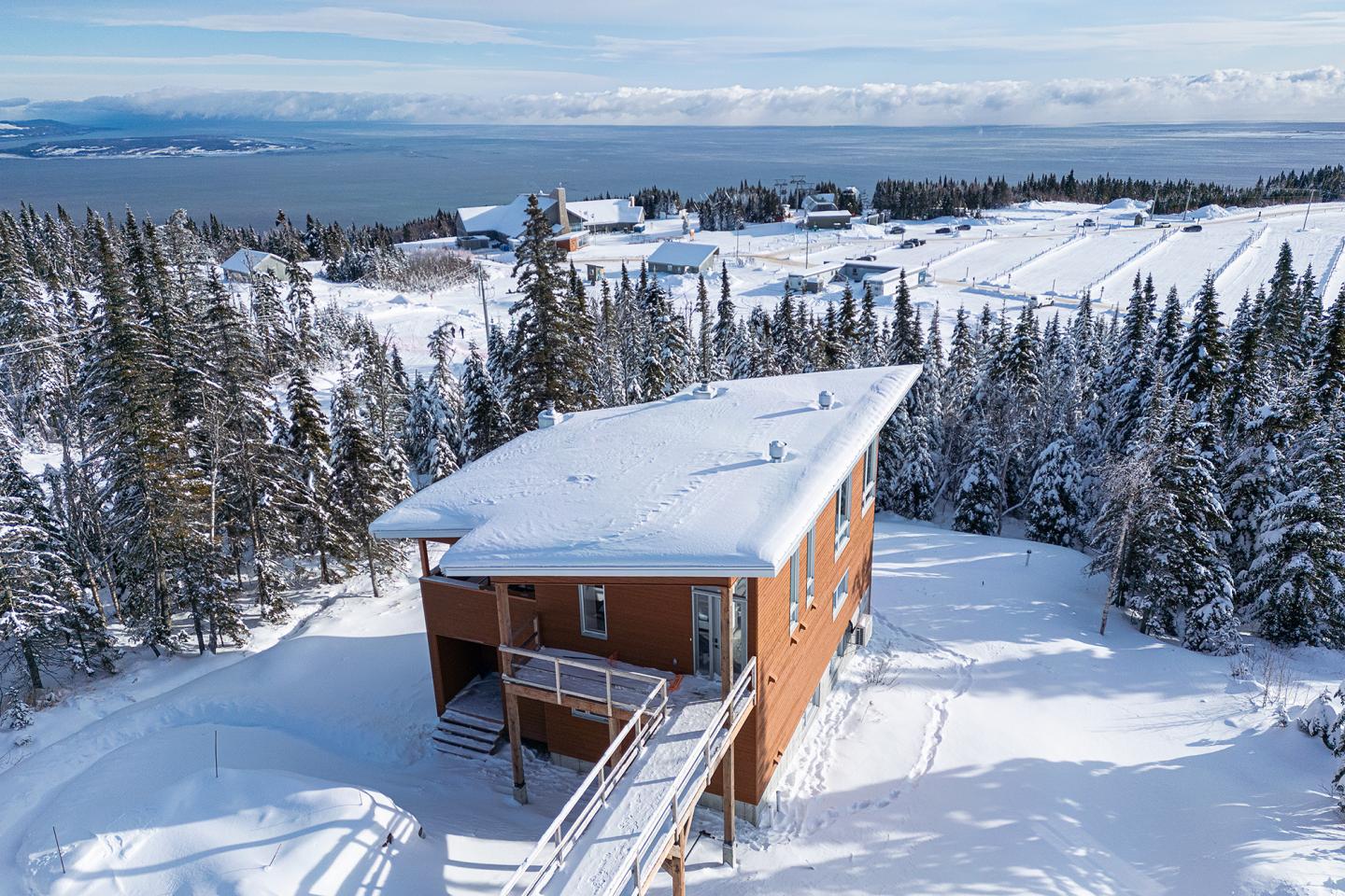 Maison en bois entourée de neige, forêt et vue sur la mer au loin.