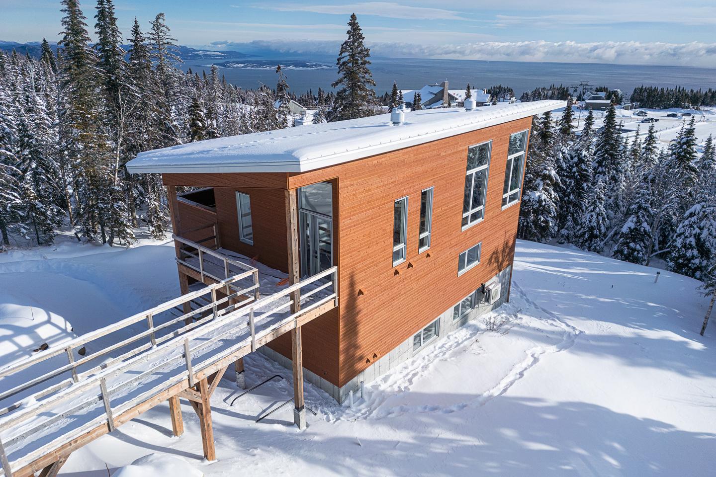 Maison en bois moderne dans un paysage enneigé avec des arbres et une vue sur la mer.