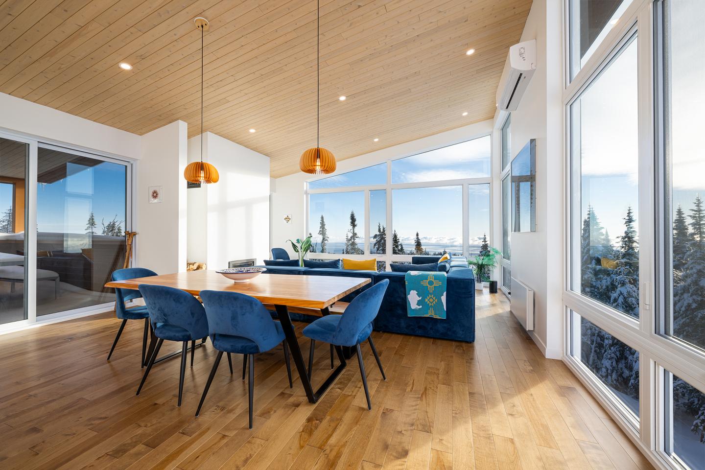 Salle à manger lumineuse avec table en bois, chaises bleues, grandes fenêtres et vue sur forêt.