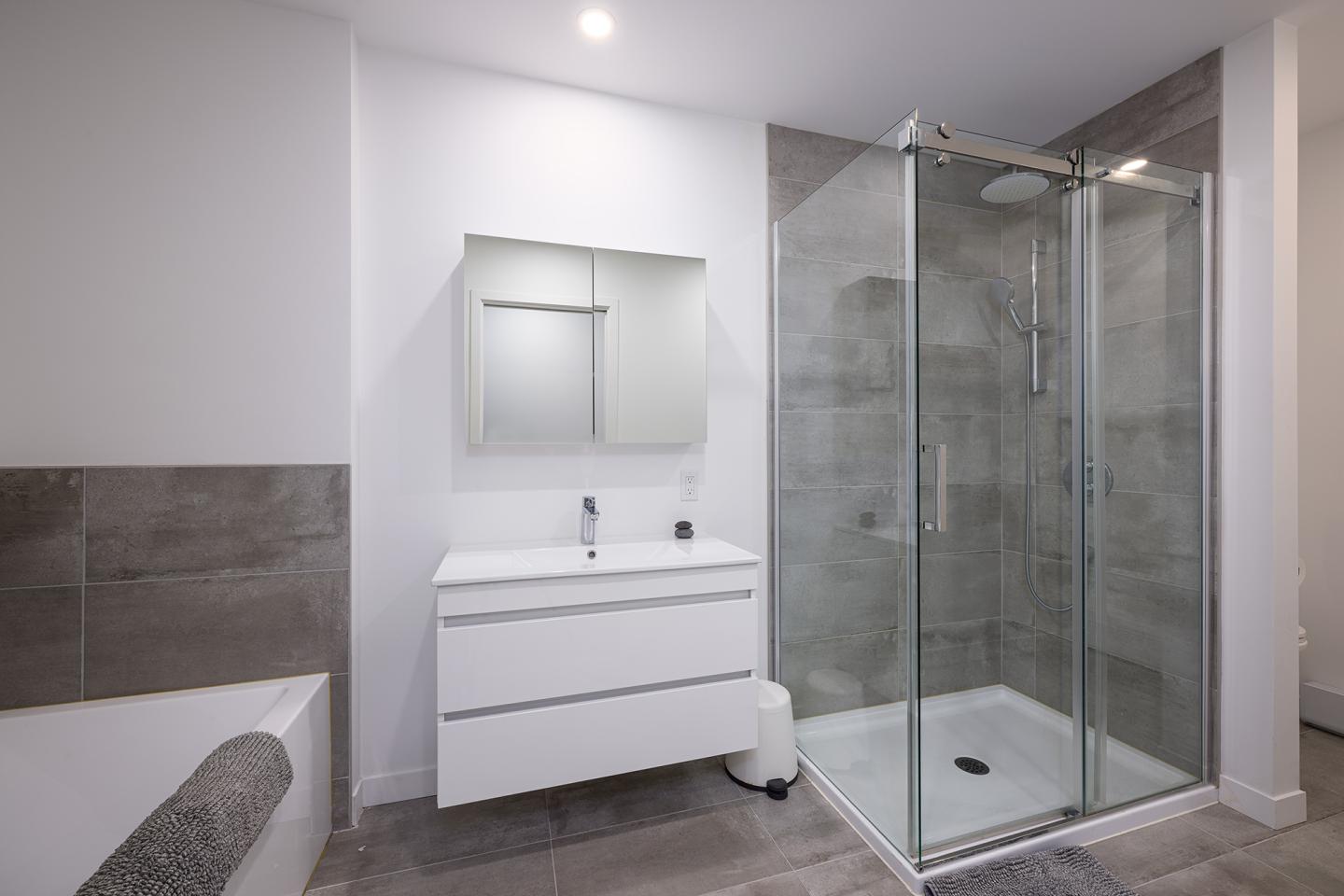 Salle de bain moderne avec douche en verre, lavabo blanc et mur gris.