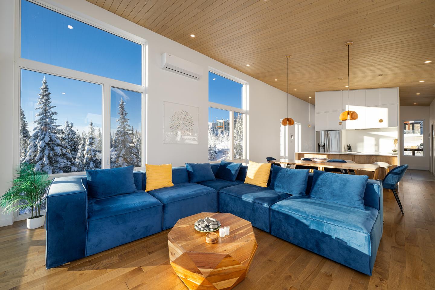 Salon moderne avec canapé bleu, coussins jaunes, et grandes fenêtres donnant sur des arbres enneigés.