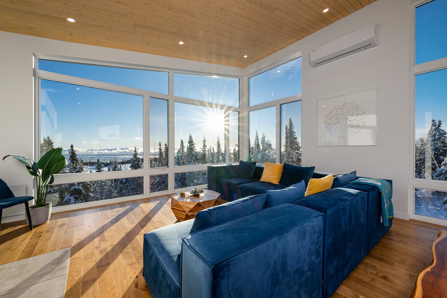 Salon moderne avec canapés bleus, grandes fenêtres et vue sur la forêt enneigée.