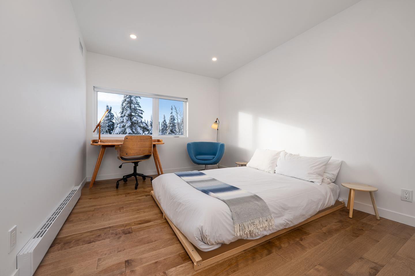 Chambre minimaliste avec lit double, bureau près de la fenêtre, arbres visibles dehors.