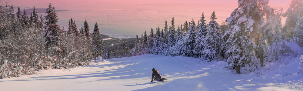 Personne sur une piste enneigée entourée de sapins, ciel rose au crépuscule.