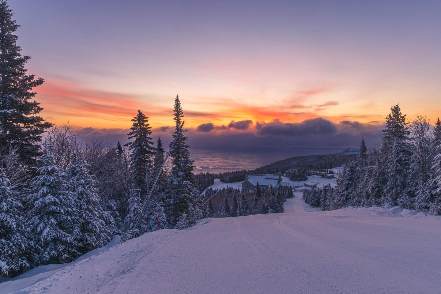 Piste enneigée au coucher de soleil, arbres et ciel coloré.