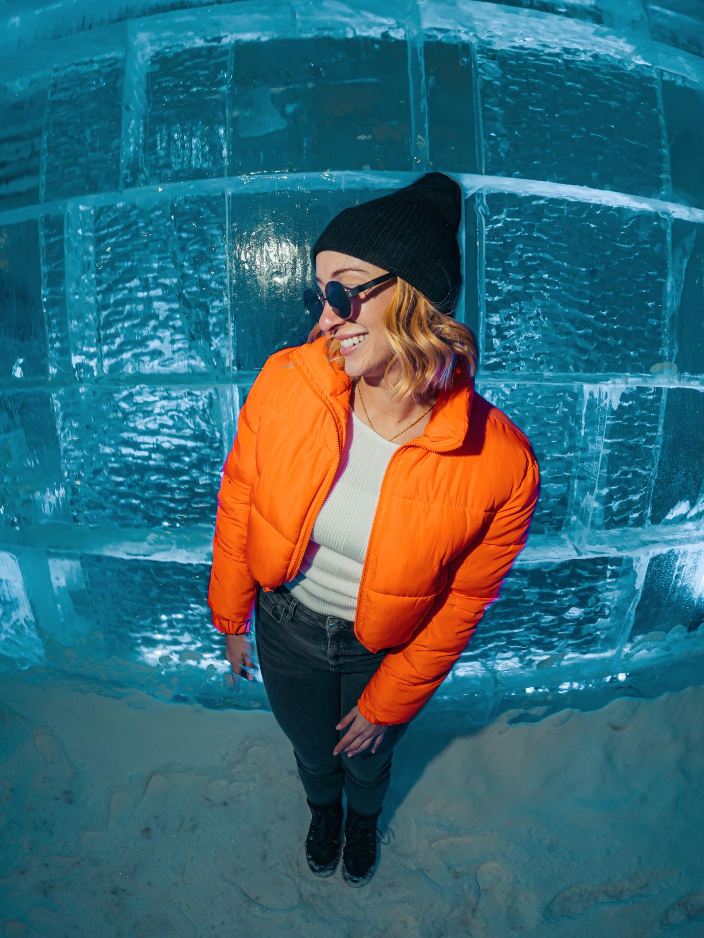 Personne souriante portant veste orange et bonnet noir devant un mur de glace bleutée.