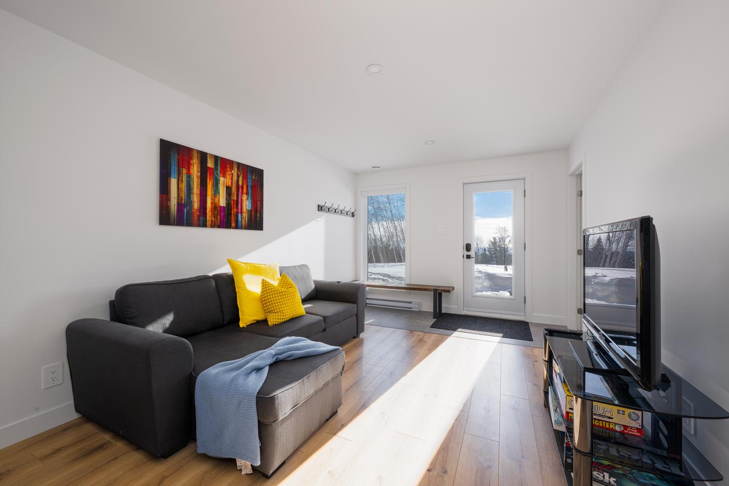 Salon moderne avec canapé gris, coussins jaunes, et télévision. Lumière du jour abondante.