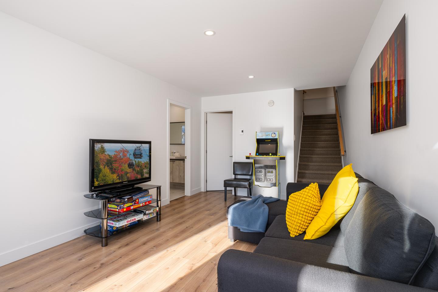 Salon moderne avec canapé gris, coussins jaunes, télé et tableau coloré. Escalier au fond.