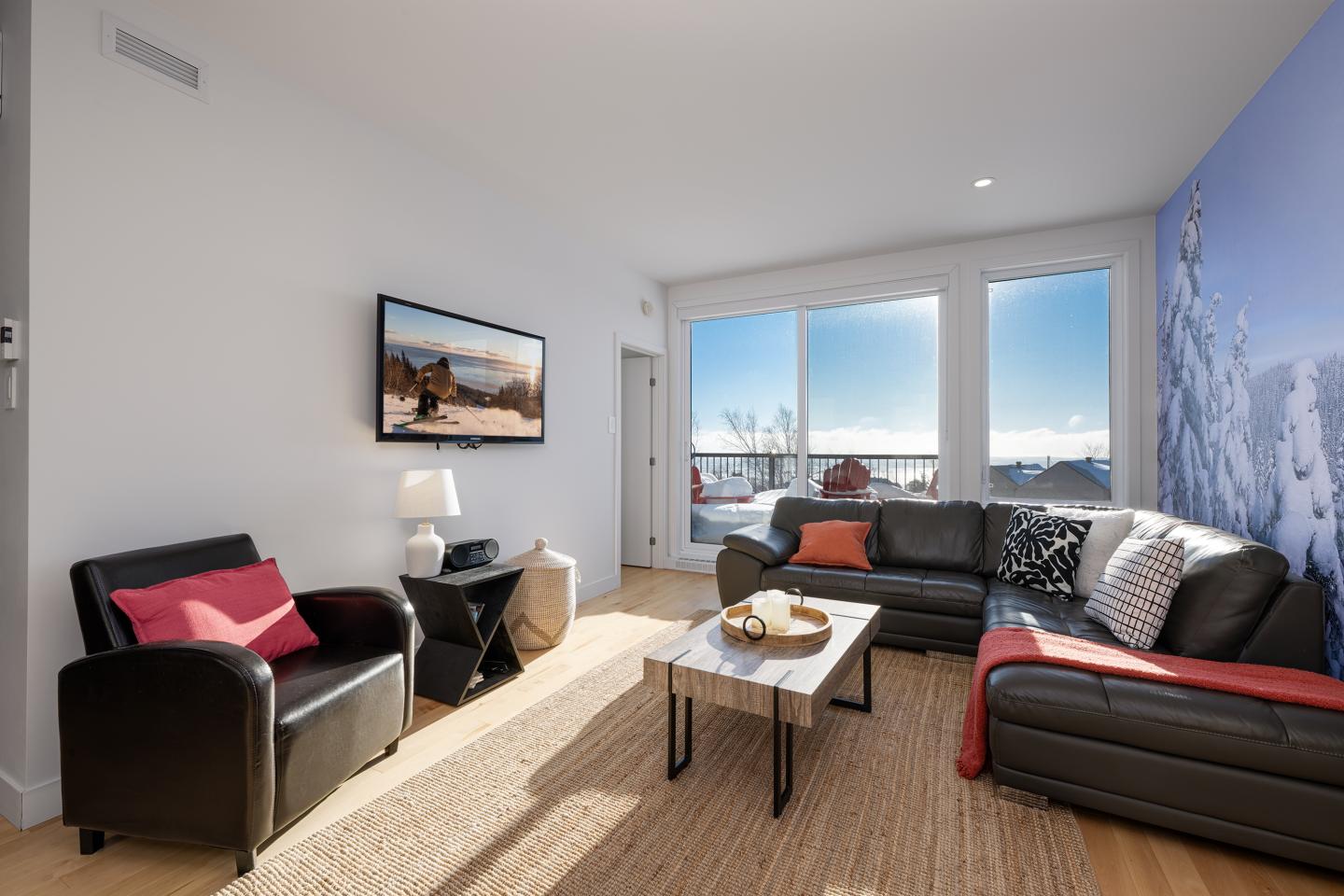 Salon moderne avec canapés noirs, grande fenêtre ensoleillée et photo murale de montagne.