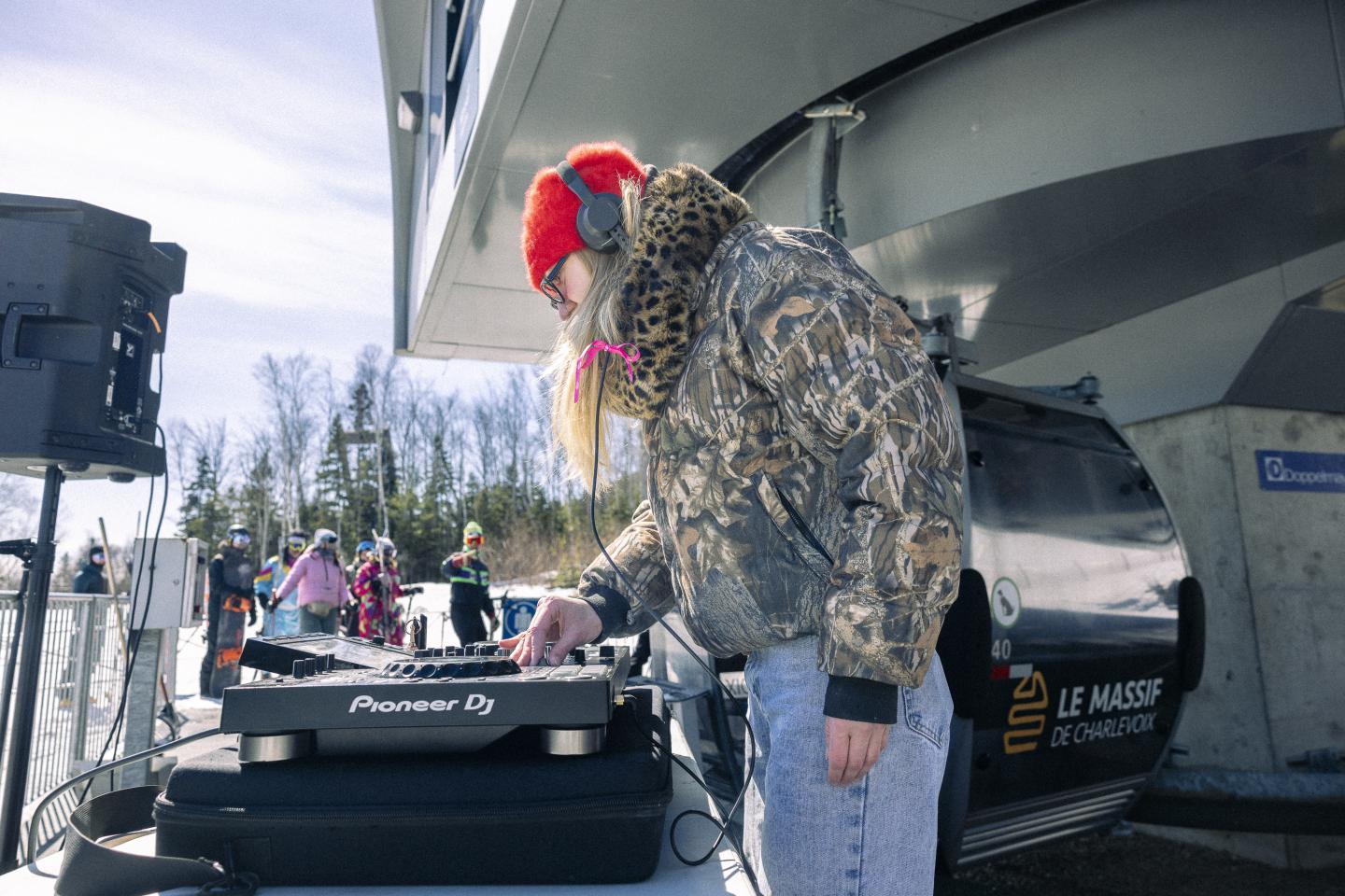 DJ avec casquette rouge mixant en plein air, près d'une station de ski.