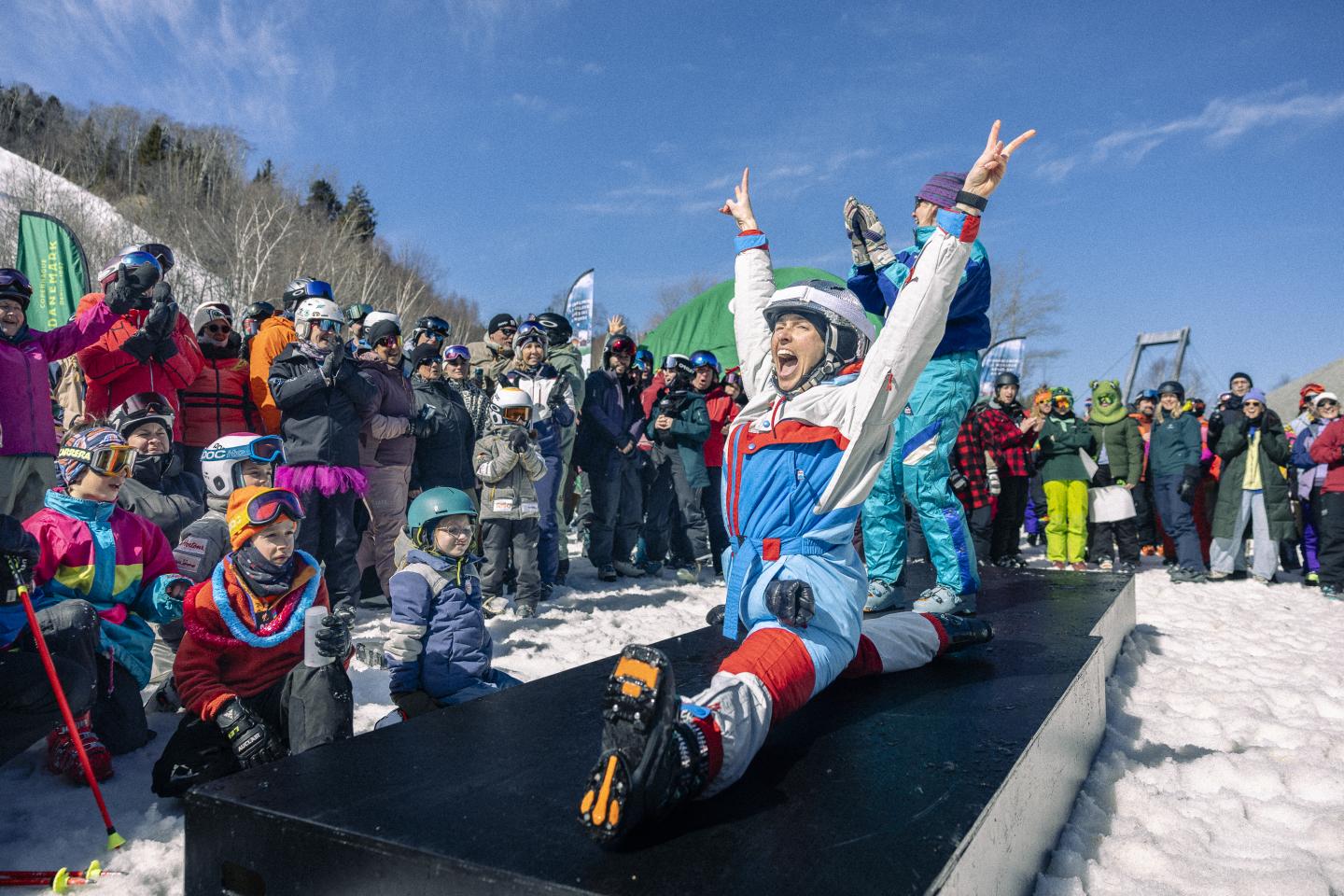 Personne vêtue de façon colorée posant joyeusement sur une rampe enneigée sous un ciel bleu.