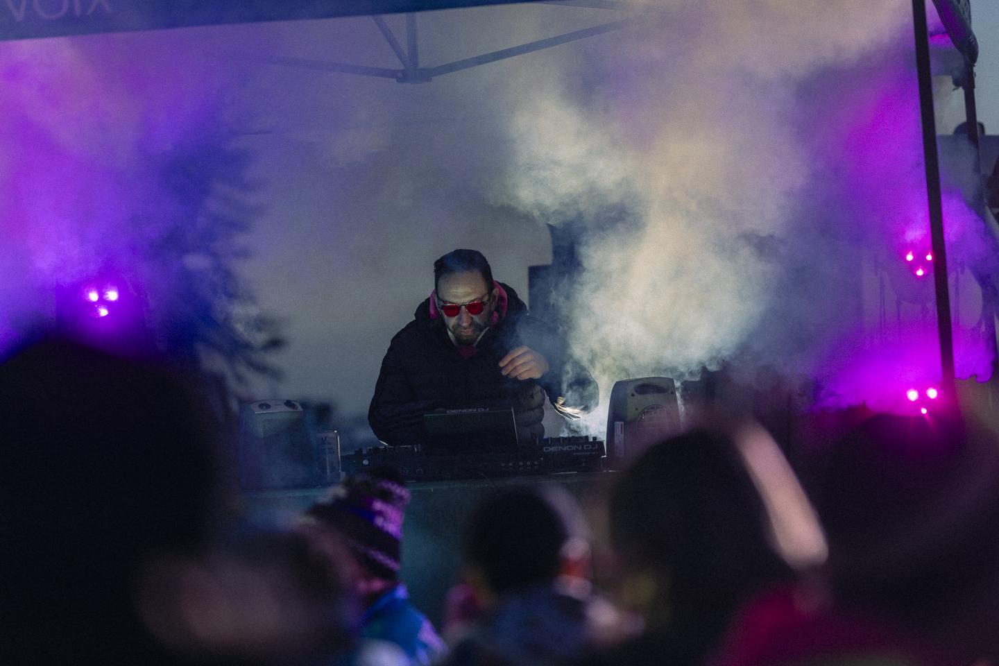 DJ en lunettes rouges mixant sur scène, fumée et lumières violettes autour.
