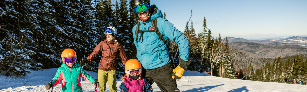 Famille en ski sur une pente enneigée, arbres et montagnes en arrière-plan.