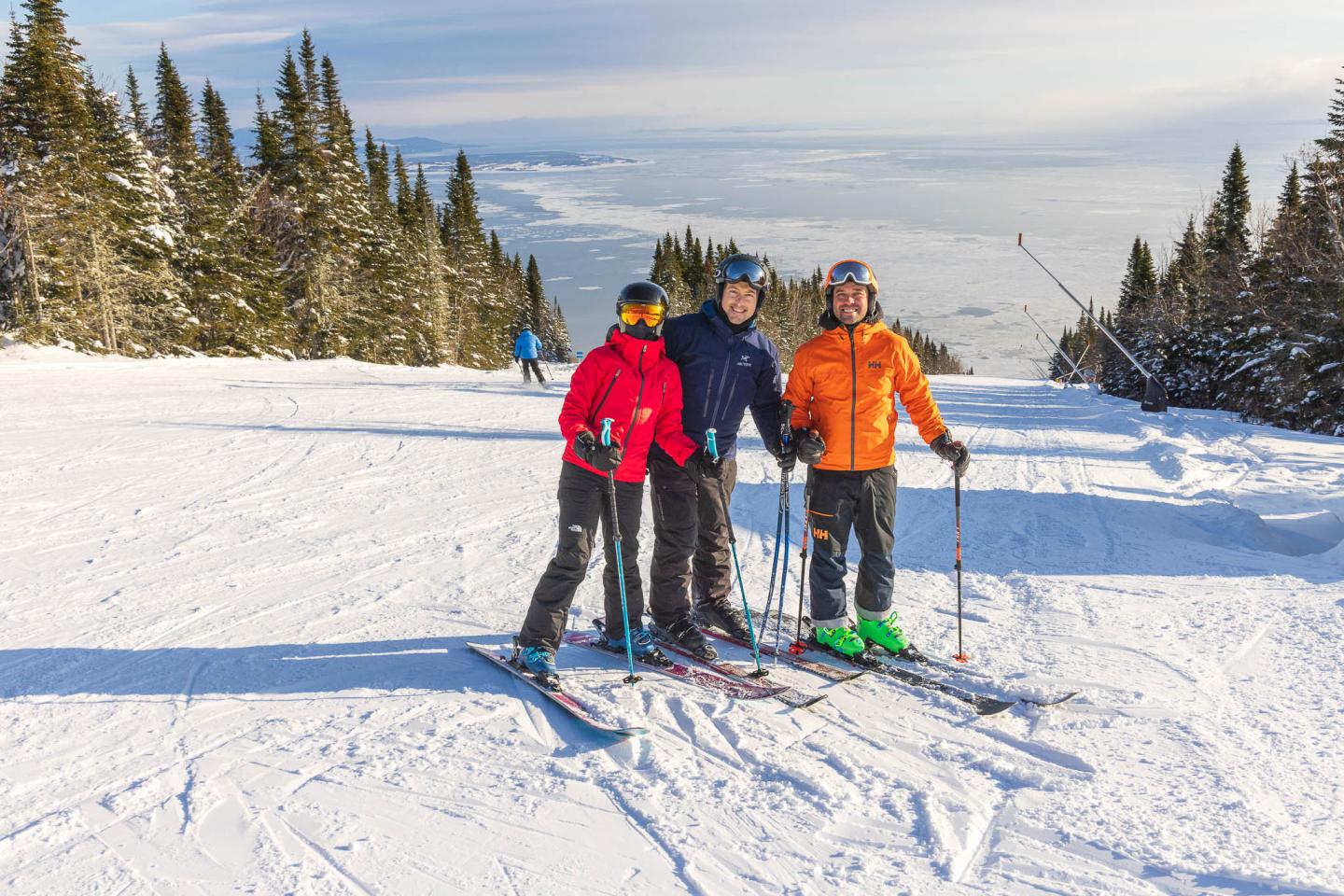 Trois personnes en ski souriantes sur une piste enneigée, ciel dégagé et arbres en arrière-plan.