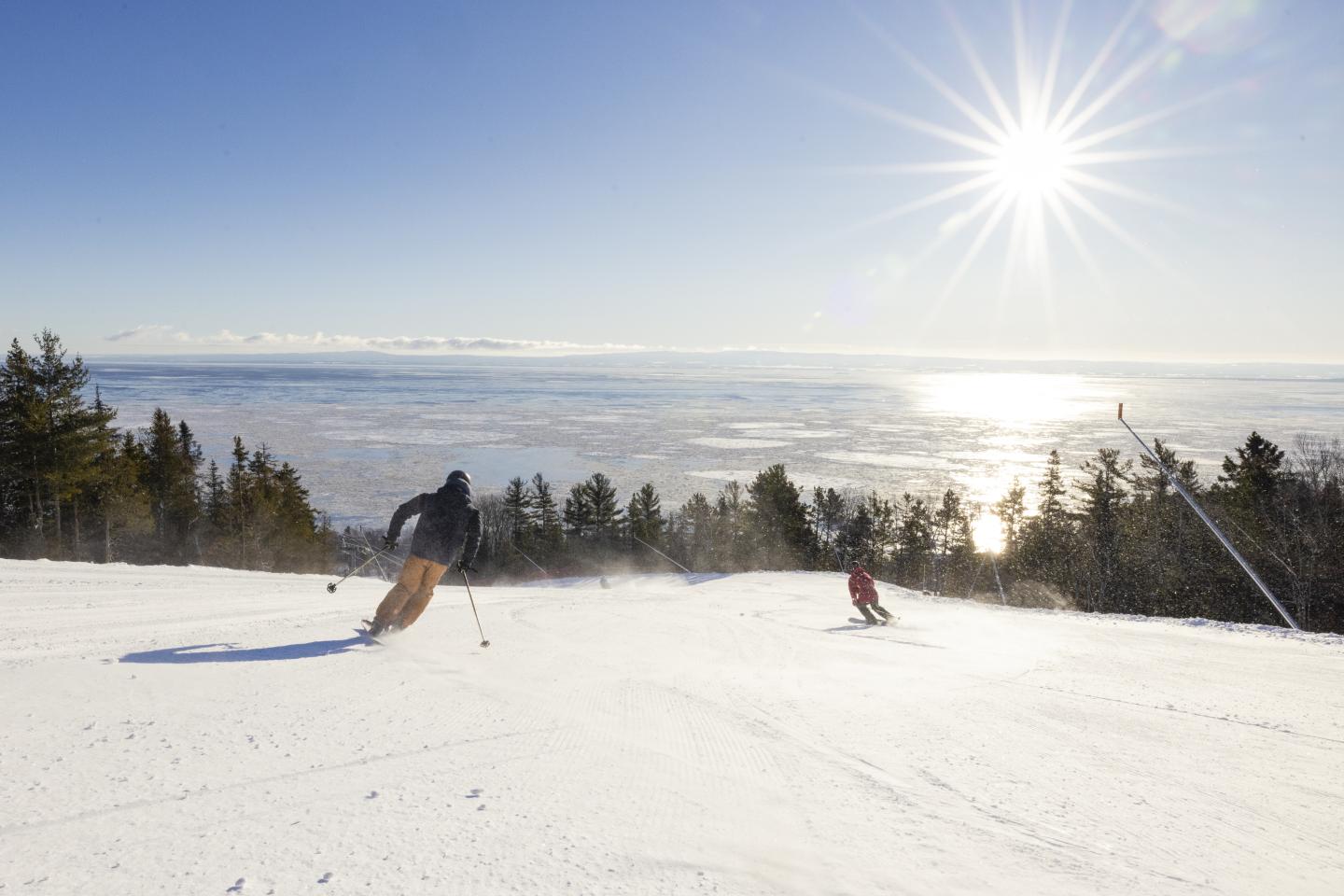 Deux skieurs descendent une piste enneigée sous un soleil éclatant, vue sur paysage hivernal.