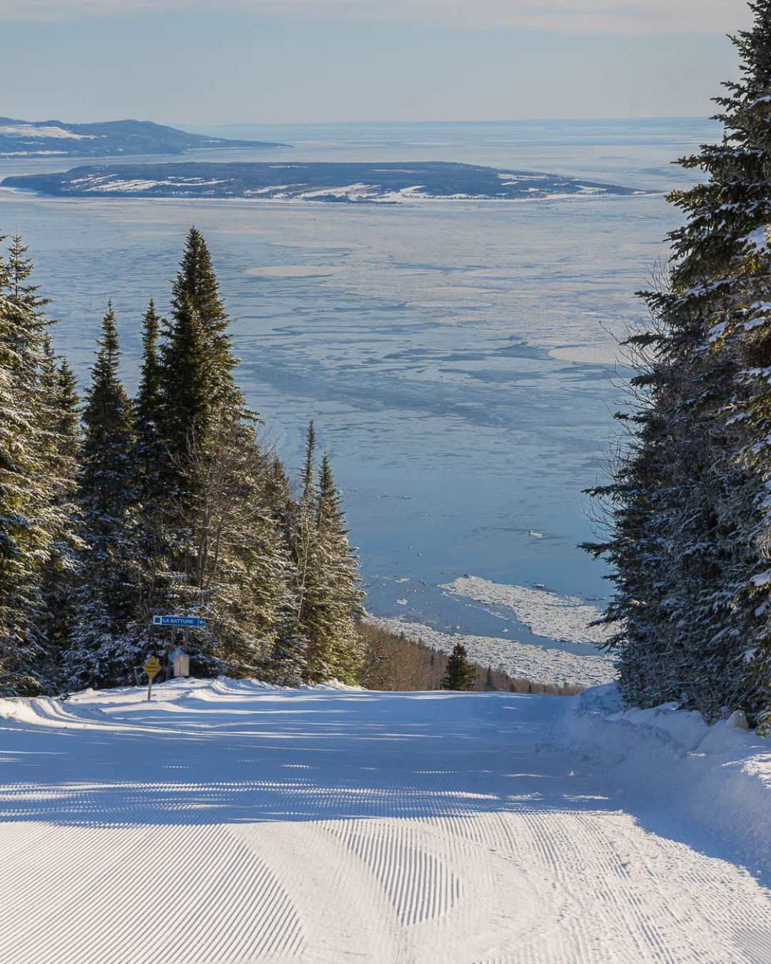 Piste de ski enneigée entourée de sapins, vue sur un lac gelé en arrière-plan.