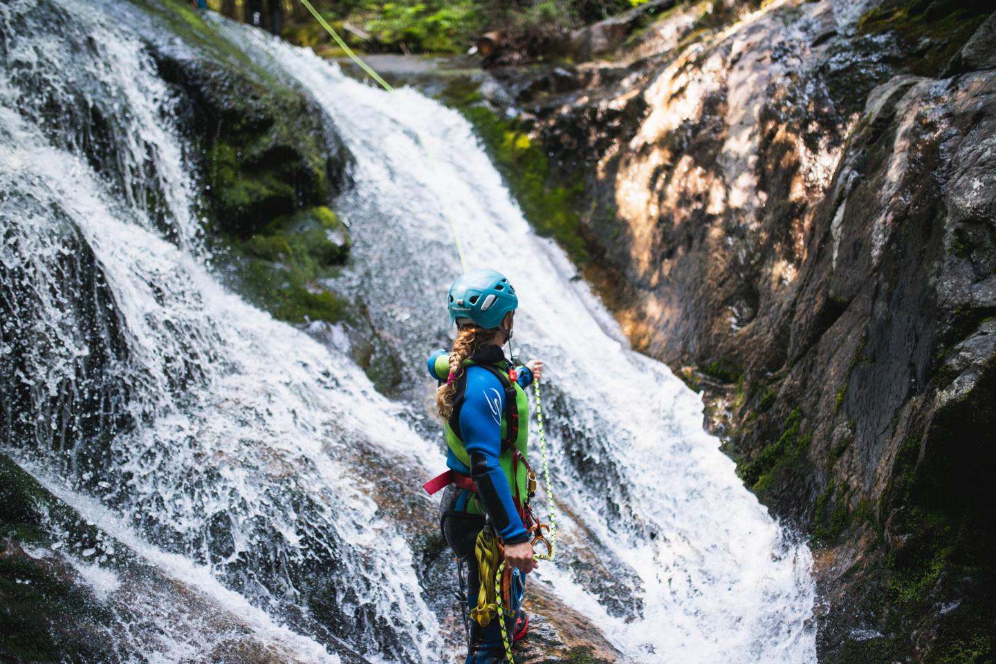 Personne en équipement escalade devant une cascade, dans une forêt lumineuse.