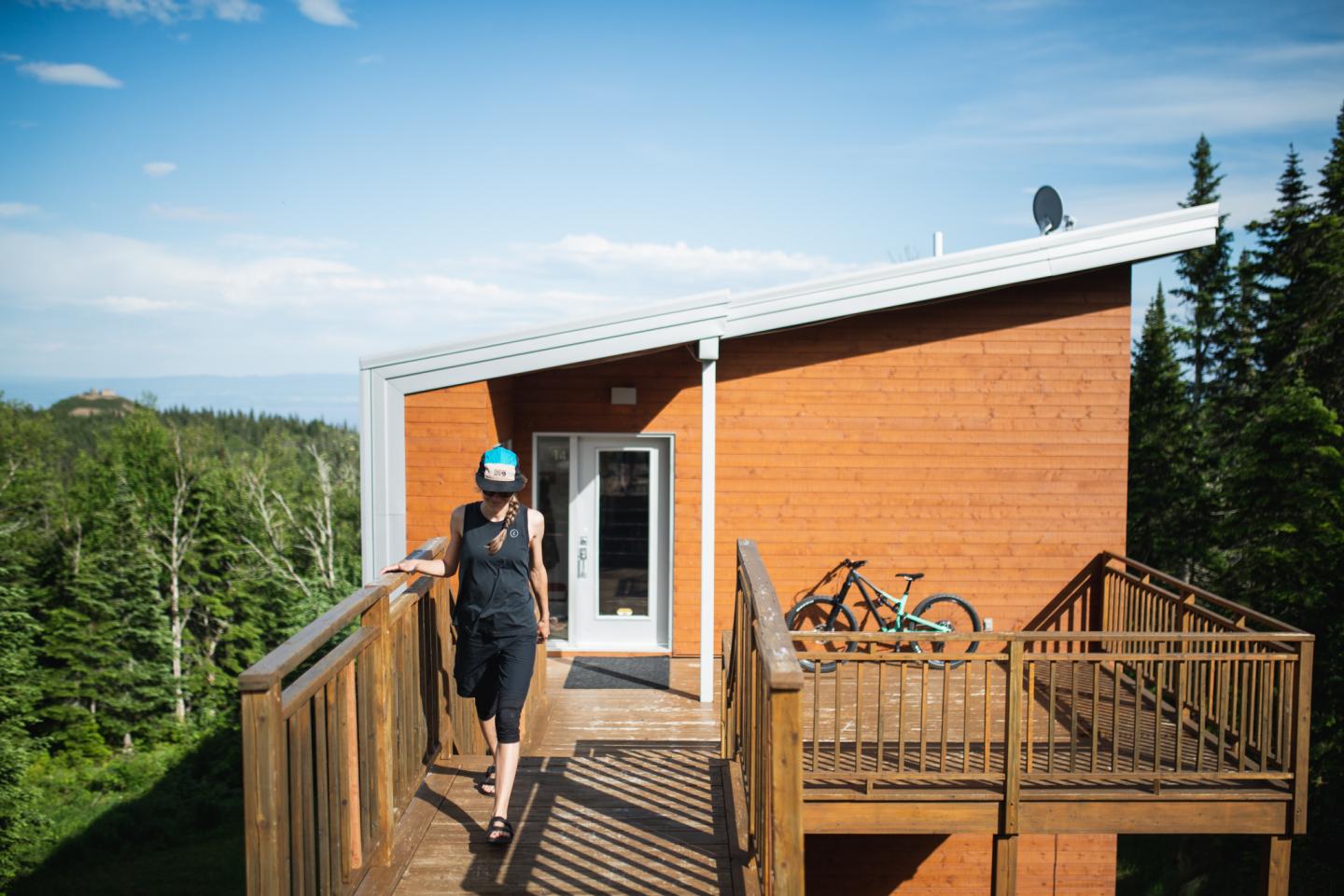 Personne avec casque devant une maison en bois, vélos sur le balcon, ciel bleu.